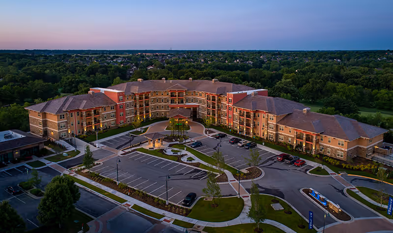 Aerial view of Fairways of Ironhorse Independent Living facility at dusk, showing a large, multi-story building with a curved design surrounded by parking lots, landscaped areas, and trees in the background.
