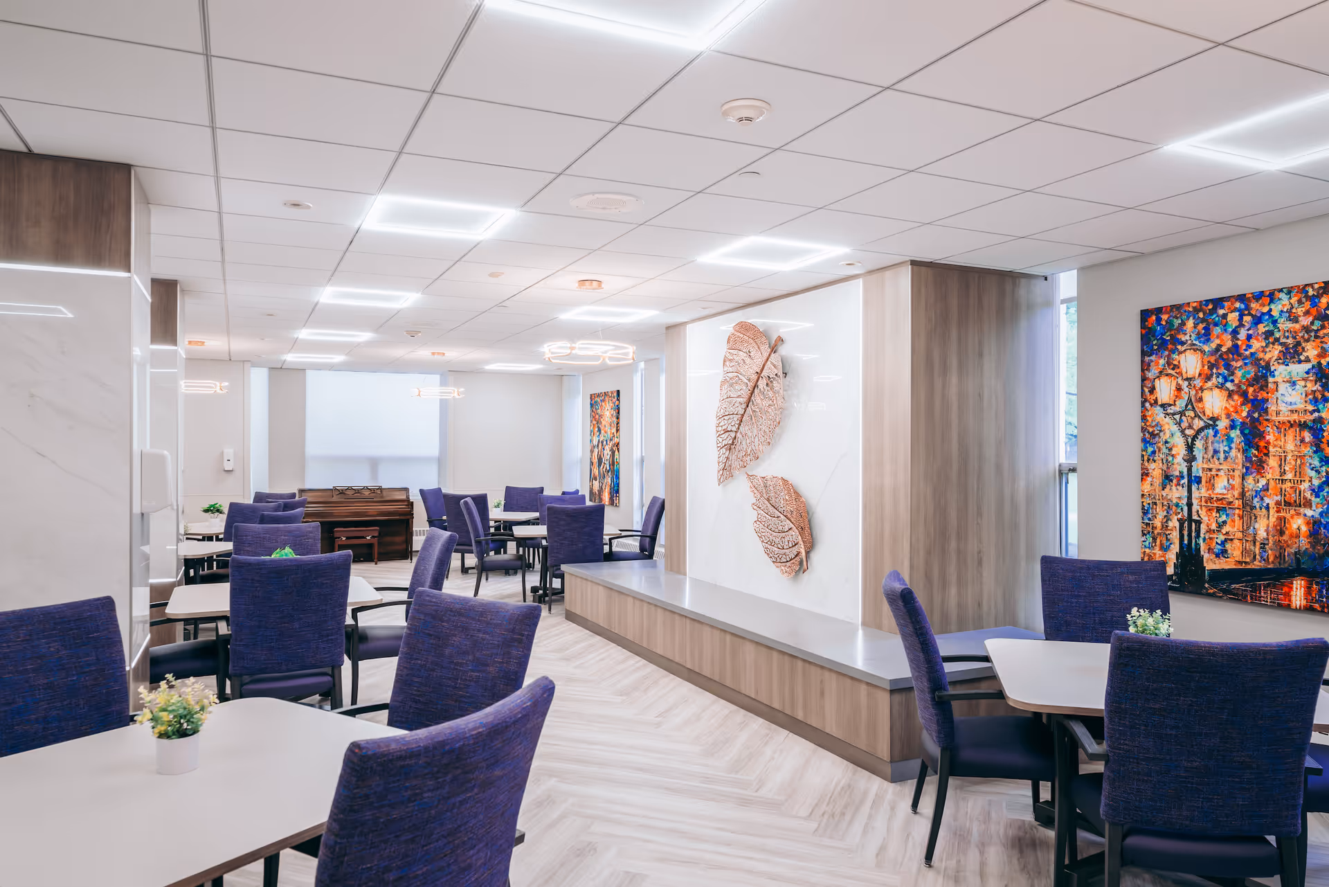 A modern dining room with multiple tables and purple upholstered chairs. The room features light wood flooring in a herringbone pattern, white walls, and ceiling with recessed lighting. Decorative elements include two large metallic leaf wall sculptures and a colorful painting of a cityscape with street lamps. A piano is visible in the background.