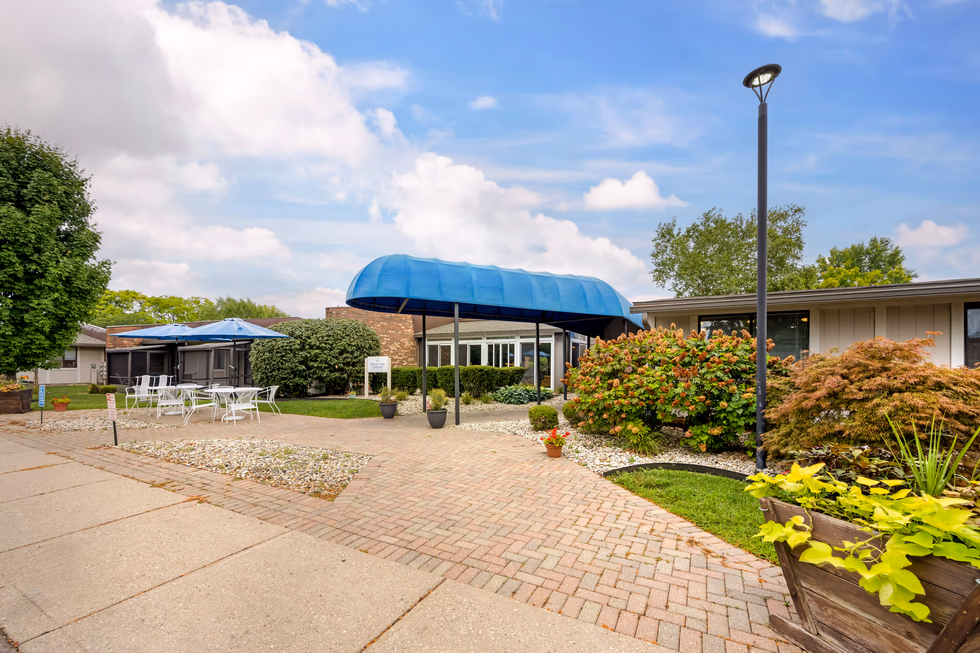 Outdoor entrance area of a senior living facility with a blue canopy over the walkway, surrounded by bushes and plants. There are white metal tables and chairs with blue umbrellas on a paved patio to the left, and a tall street lamp on the right. The sky is partly cloudy.