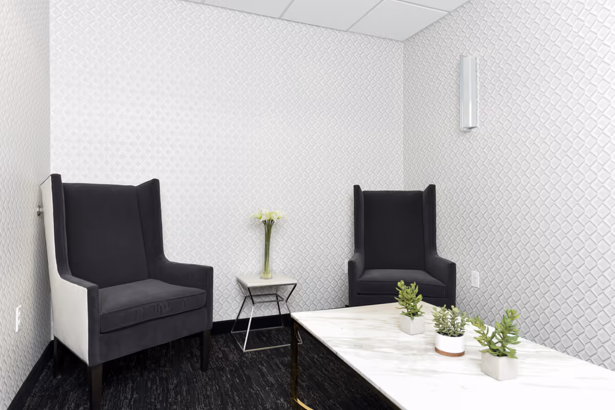 A small, modern sitting area with two black and white armchairs, a white marble coffee table with three small potted plants, and a small side table with a vase of white flowers. The walls have a textured white geometric pattern, and the floor is covered with dark carpet.