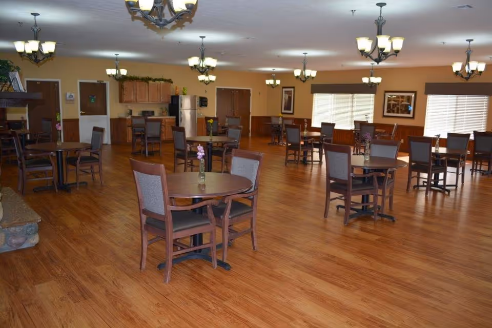 A spacious dining room with multiple round wooden tables and chairs arranged neatly on a wooden floor. Each table has a small vase with flowers. The room has warm beige walls, several windows with blinds, framed pictures on the walls, and multiple ceiling light fixtures. In the background, there is a kitchen area with cabinets, a refrigerator, and a countertop.