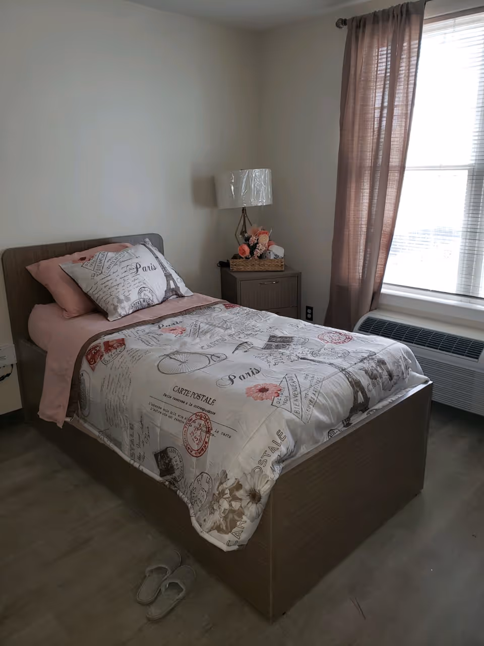 A small bedroom with a single bed featuring Paris-themed bedding and pink pillows. Next to the bed is a wooden nightstand with a lamp and a basket of flowers. A window with sheer brown curtains allows natural light into the room. A pair of slippers is placed on the floor beside the bed.