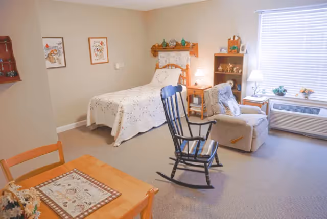 A cozy senior living bedroom with a single bed covered in a white floral quilt, a wooden rocking chair in the center, a cushioned recliner chair near a window with blinds, a wooden nightstand with a lamp, and a small wooden bookshelf with decorative items. A wooden table with two chairs and a decorative placemat is partially visible in the foreground.