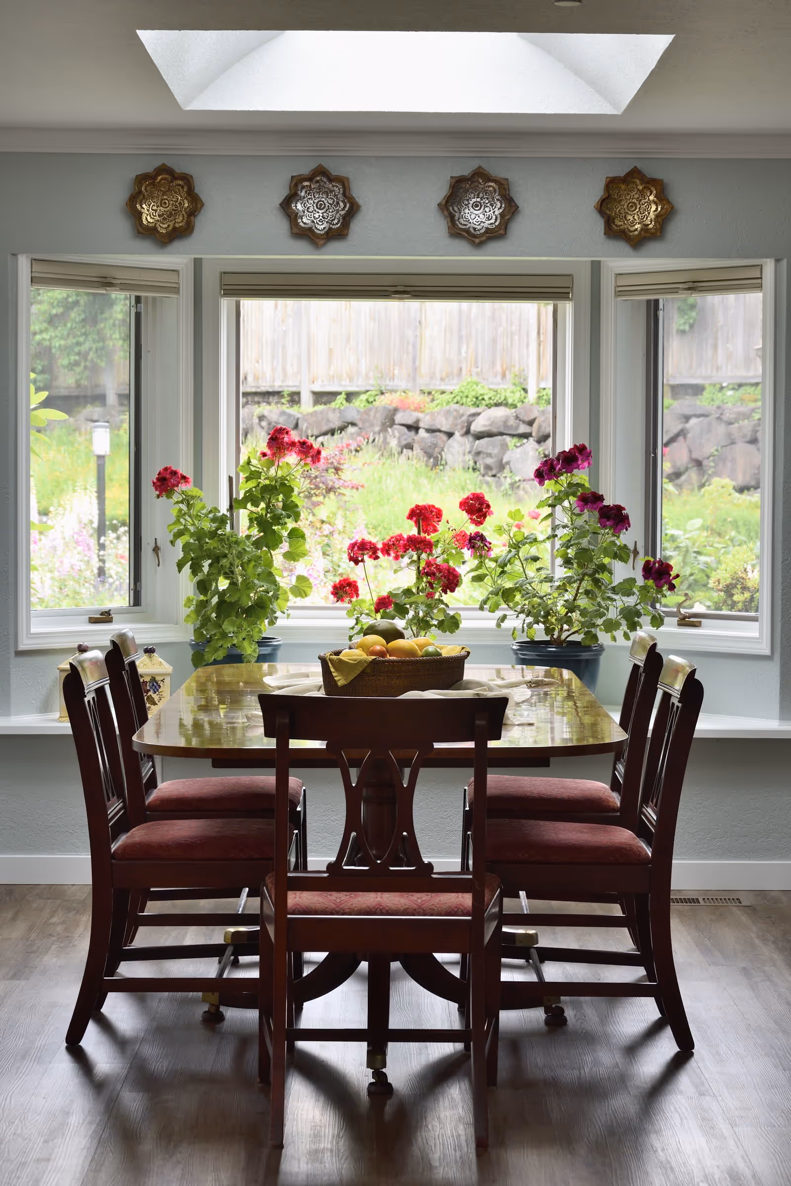 A dining area with a wooden table and six chairs with red cushions. On the table is a basket of fruit. Behind the table is a large bay window with three sections, showing a garden with green plants and flowers outside. There are four decorative wall hangings above the window and a skylight in the ceiling.
