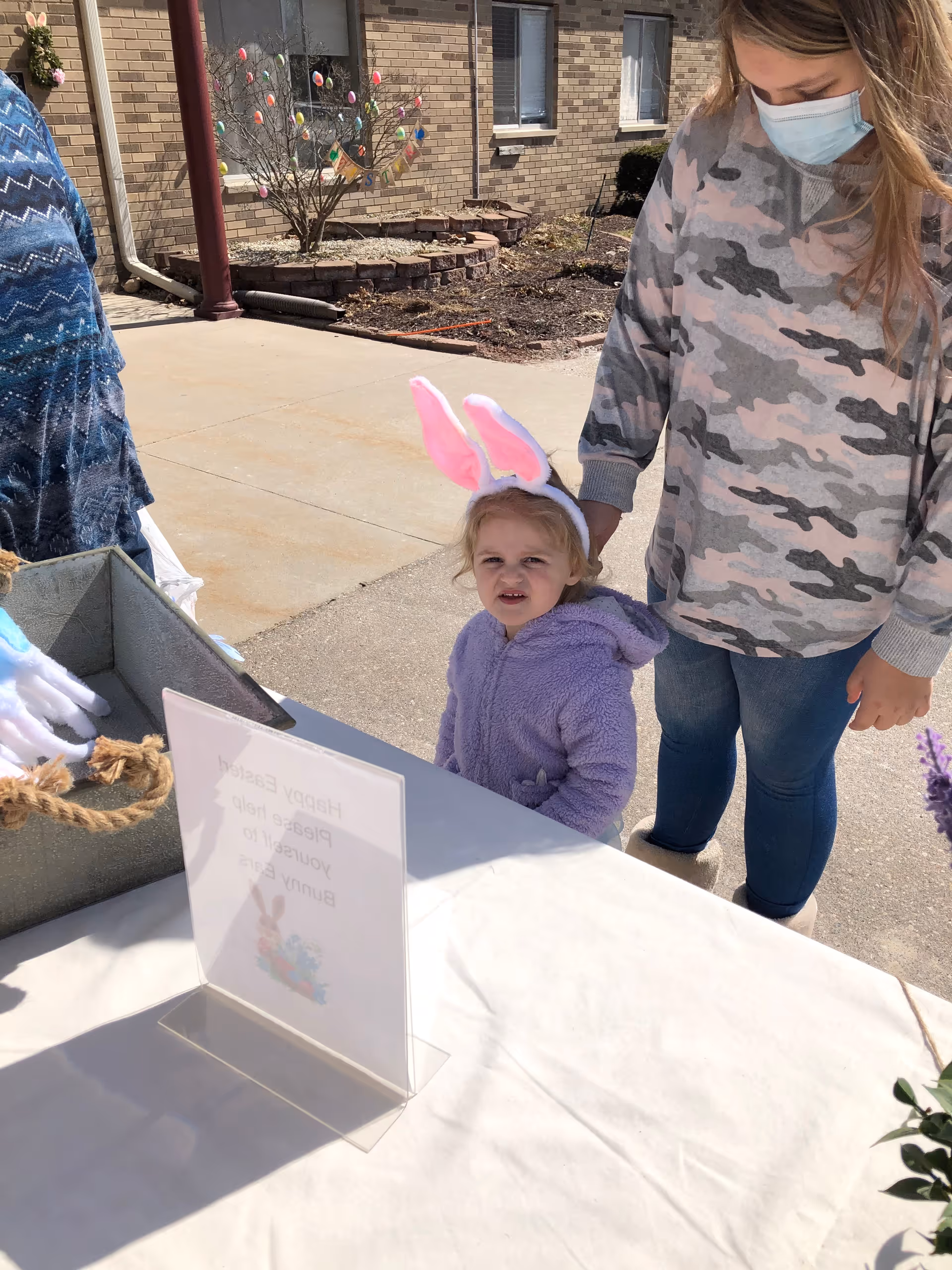 A young child wearing pink bunny ears and a purple jacket stands next to an adult wearing a camouflage sweatshirt and a face mask outside a brick building. There is a table with a white tablecloth and a sign that reads 'Happy Easter Please help yourself to bunny ears.' In the background, a small tree decorated with colorful Easter eggs is visible.