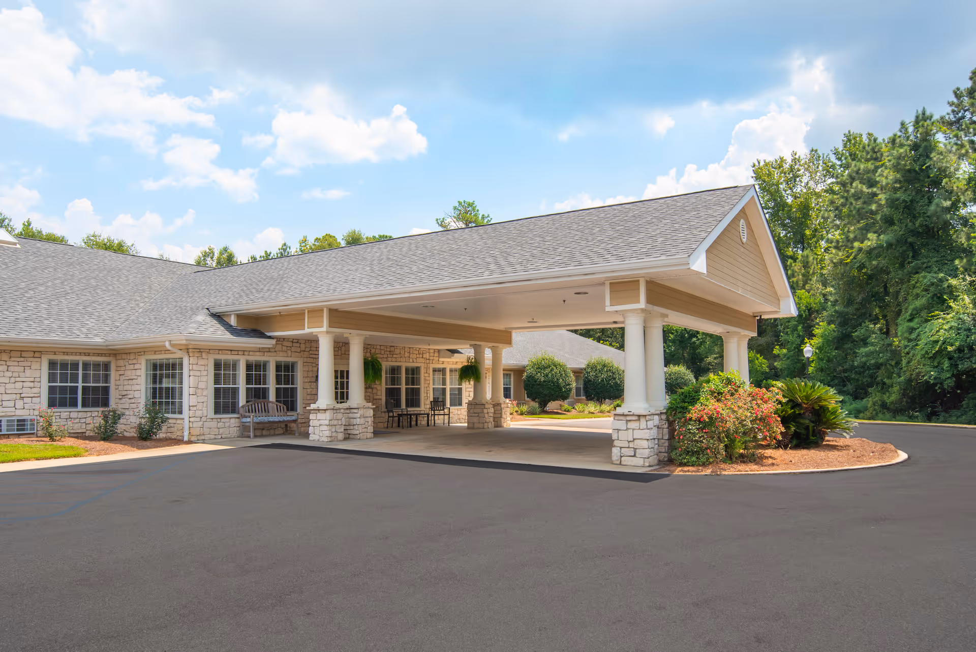 Exterior view of Brookdale Hattiesburg senior living facility showing a covered entrance with stone pillars, a bench, and surrounding greenery under a partly cloudy sky.