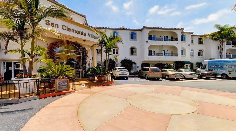 Exterior view of San Clemente Villas, a multi-story residential building with white walls and balconies. There are several palm trees and plants near the entrance, which has a sign above it reading 'San Clemente Villas'. Several cars are parked in front of the building, and a shuttle van with the facility's name is visible on the right side. The sky is partly cloudy.
