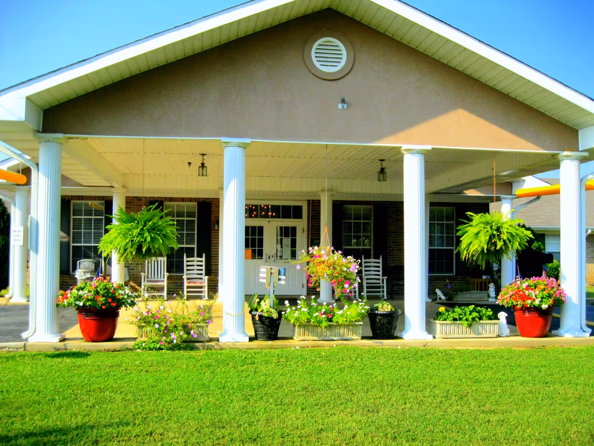 Front porch of a building with white columns, hanging ferns, potted flowers, and white rocking chairs. The building has a beige gable roof and brick walls with windows and a white door. There is a well-maintained green lawn in the foreground.