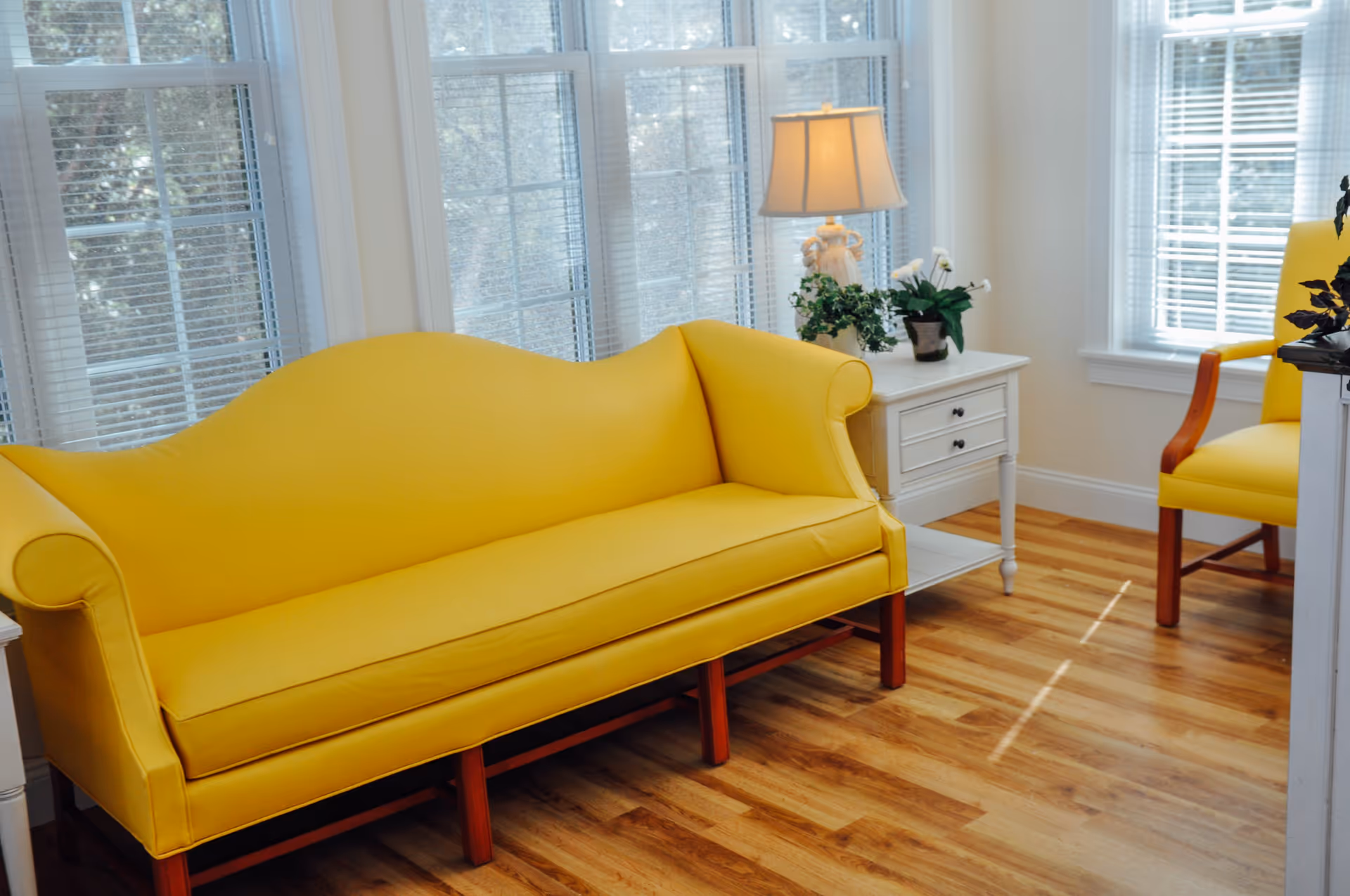Bright interior seating area with a yellow sofa and matching chair beside a white side table and lamp in front of tall windows.