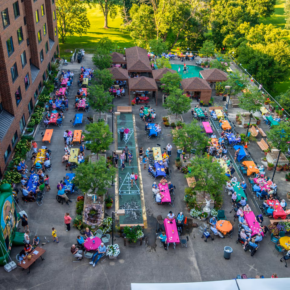 Aerial view of an outdoor gathering at Saint Therese Senior Living of New Hope, showing numerous seniors seated at colorful tables arranged around a courtyard with trees, shuffleboard courts, and small gazebo structures. The scene is lively with people socializing and enjoying the sunny day.