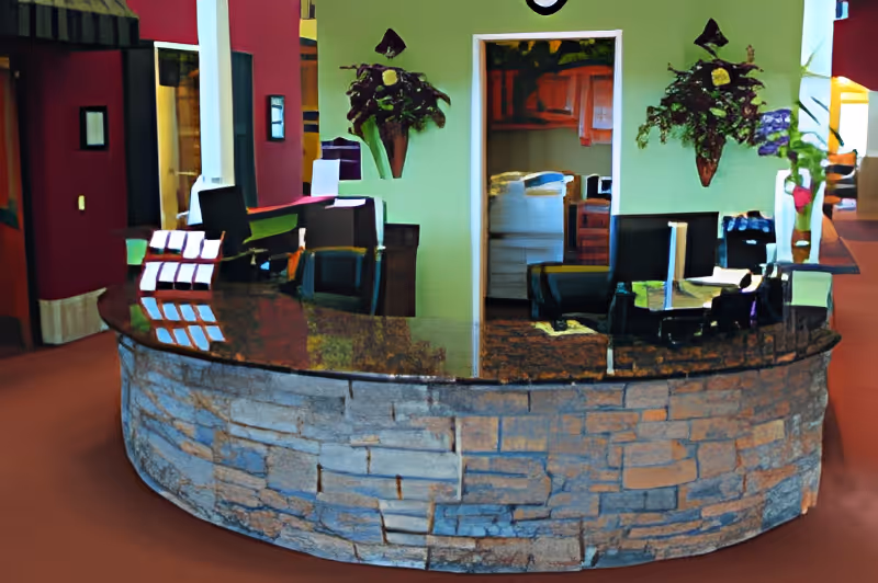 Reception desk area with a curved stone front and a dark granite countertop. Behind the desk are two black chairs and a doorway leading to another room with wooden cabinets. The walls are painted green and decorated with two wall-mounted floral arrangements. The surrounding area has red walls and carpeted flooring.