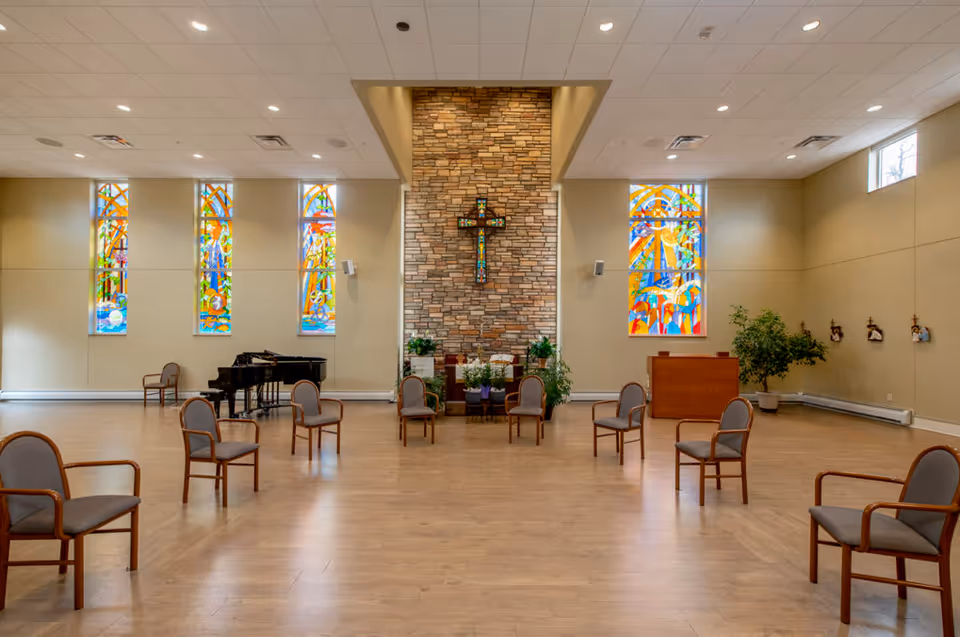 Large chapel-style interior with spaced chairs, stained-glass windows, a stone wall cross, and a grand piano.