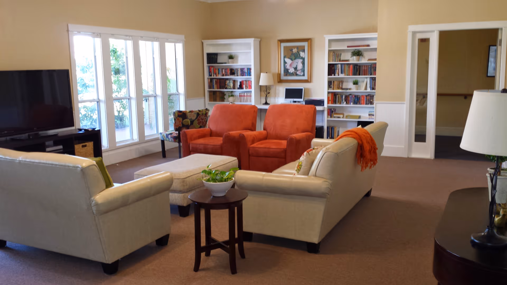 A cozy living room area in an assisted living facility featuring two beige sofas, two orange armchairs, a small round wooden table with a plant, a large flat-screen TV, bookshelves filled with books, a computer desk with a lamp, and large windows letting in natural light.