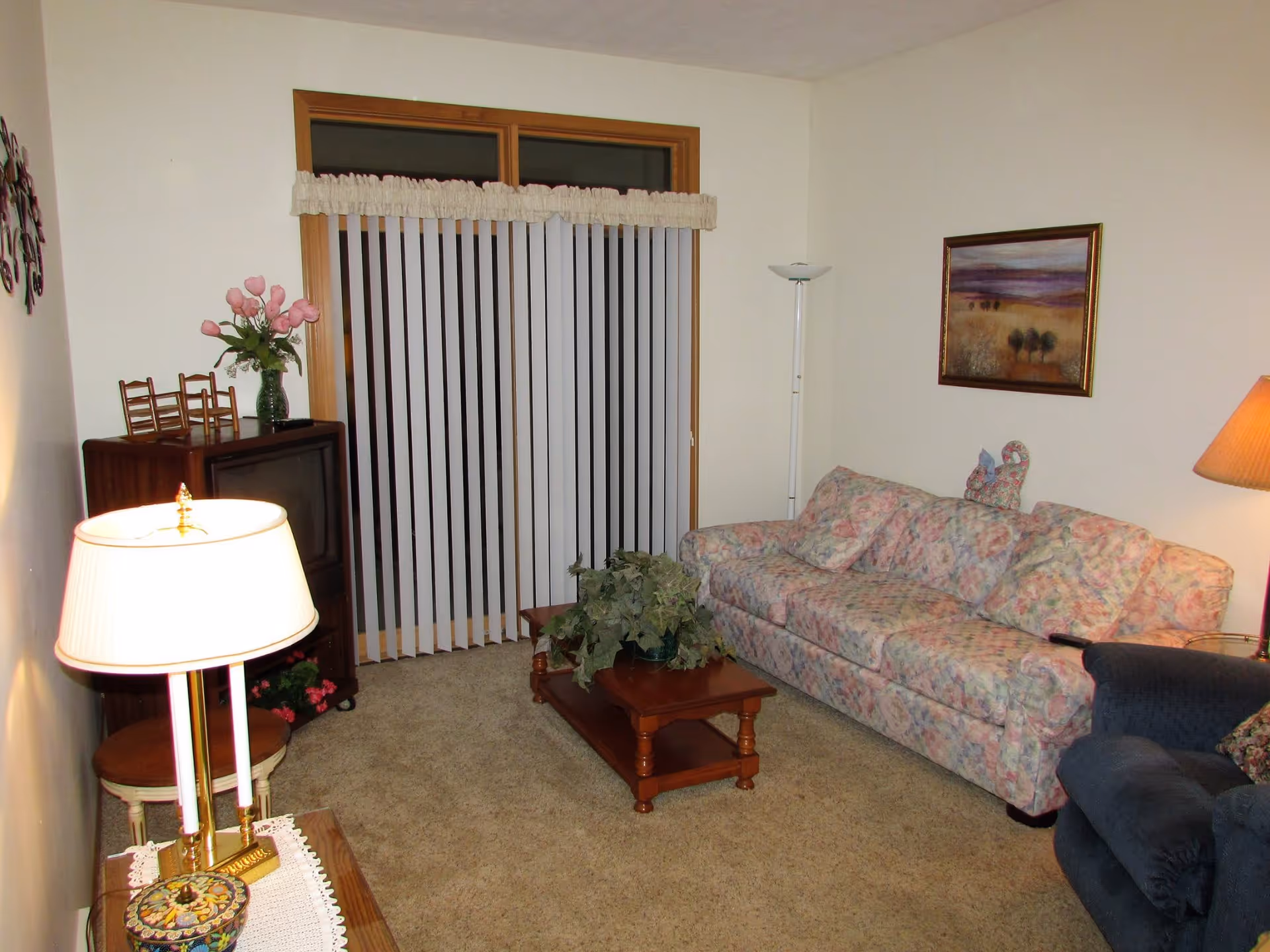 A cozy living room with a floral patterned sofa, a dark blue armchair, a wooden coffee table with a green plant, a wooden TV stand with a television and a vase of pink flowers, vertical blinds covering a sliding glass door, a floor lamp, and a table lamp on a side table.