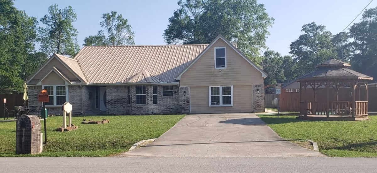 Single-story residential building with a metal roof, brick and siding exterior, a driveway leading to the front, and a wooden gazebo on the right side surrounded by green grass and trees in the background.