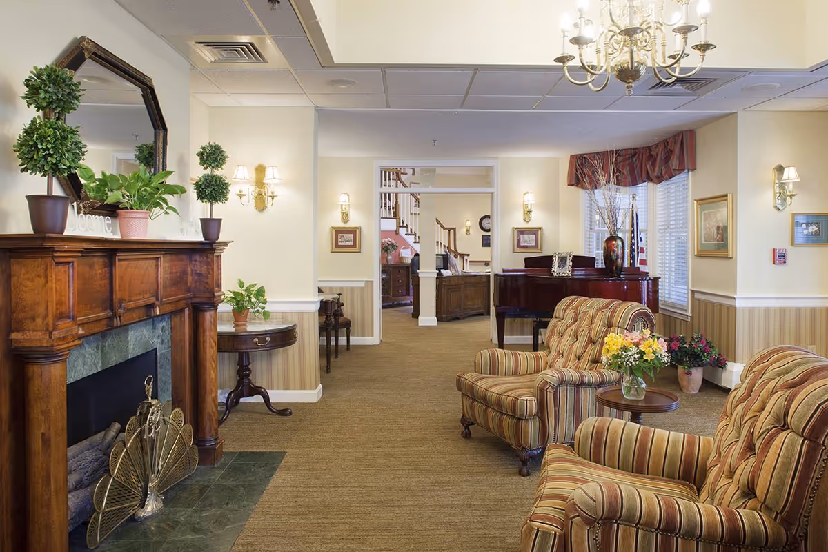 A cozy living room area in a senior living facility featuring two striped upholstered armchairs with a small round table holding a vase of flowers between them. There is a wooden fireplace mantel decorated with potted plants and a mirror above it on the left side. In the background, a grand piano with a vase and decorative items is positioned near windows with valances. The room is warmly lit with wall sconces and a chandelier, and the walls are painted light yellow with wainscoting.