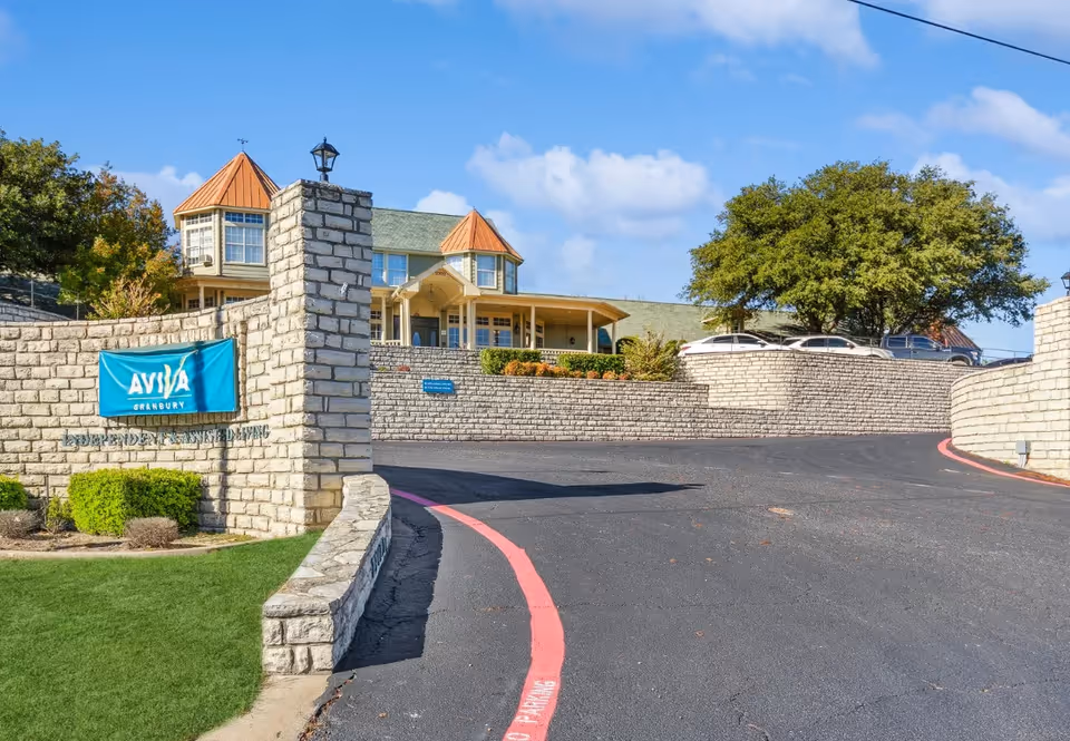 Front entrance of AVIVA Granbury Senior Living showing stone retaining walls, a blue sign, driveway, and the facility building.