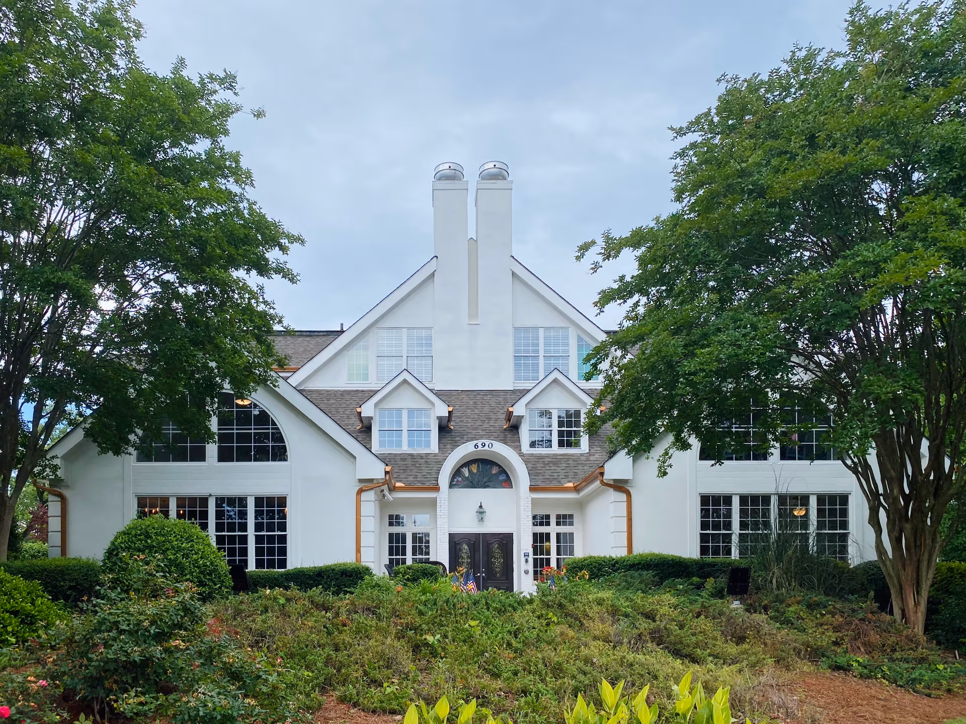 Front exterior view of a large white senior living facility building with multiple windows, two prominent chimneys, and a central entrance with double doors. The building is surrounded by green trees and bushes under a cloudy sky.