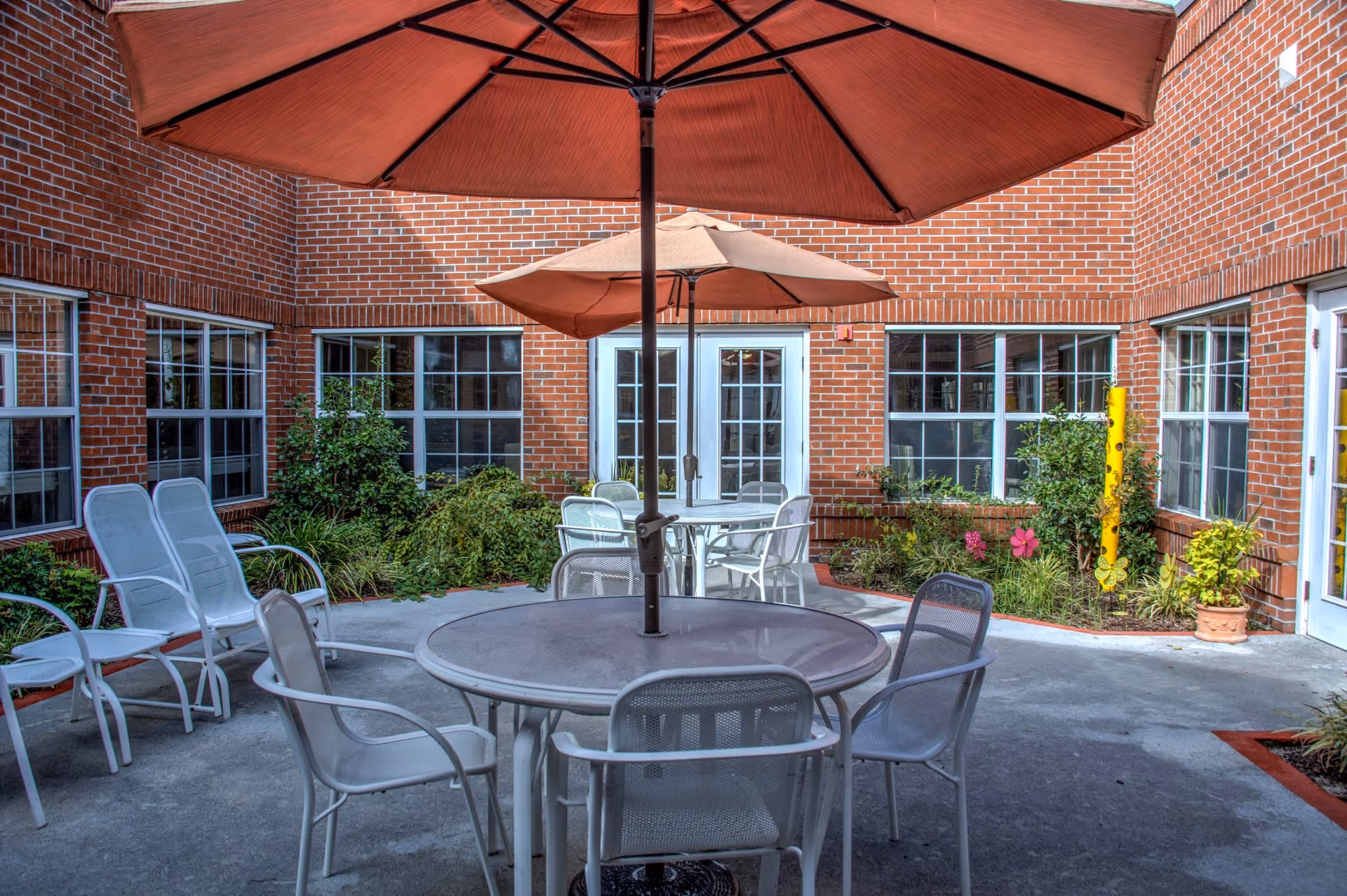 Outdoor courtyard area with round metal tables and chairs, each table shaded by a large umbrella. The courtyard is surrounded by red brick walls with multiple windows and doors. There are green plants and flowers along the edges of the courtyard.