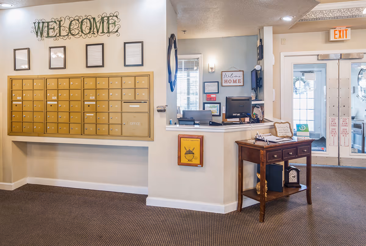 Interior view of a senior living facility lobby area with a wall of gold mailboxes labeled with numbers, a reception desk with a computer and office chair, a wooden table with drawers and decorative items, and double glass exit doors. The walls have welcoming signs including a large decorative 'WELCOME' sign above the mailboxes and a 'Welcome HOME' sign near the reception desk.