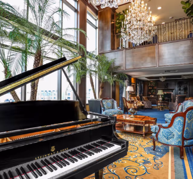 Sunlit grand lounge with a Steinway grand piano, chandeliers, palm plants, and upholstered seating.