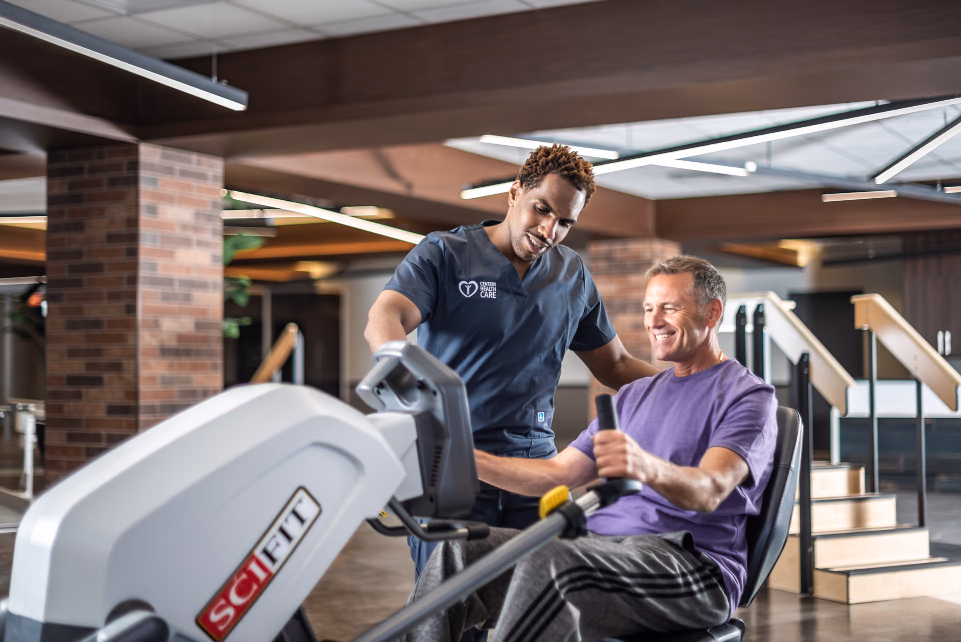 A healthcare professional in navy scrubs assists a smiling middle-aged man using a SciFit exercise machine in a modern rehabilitation facility with brick pillars and wooden accents.