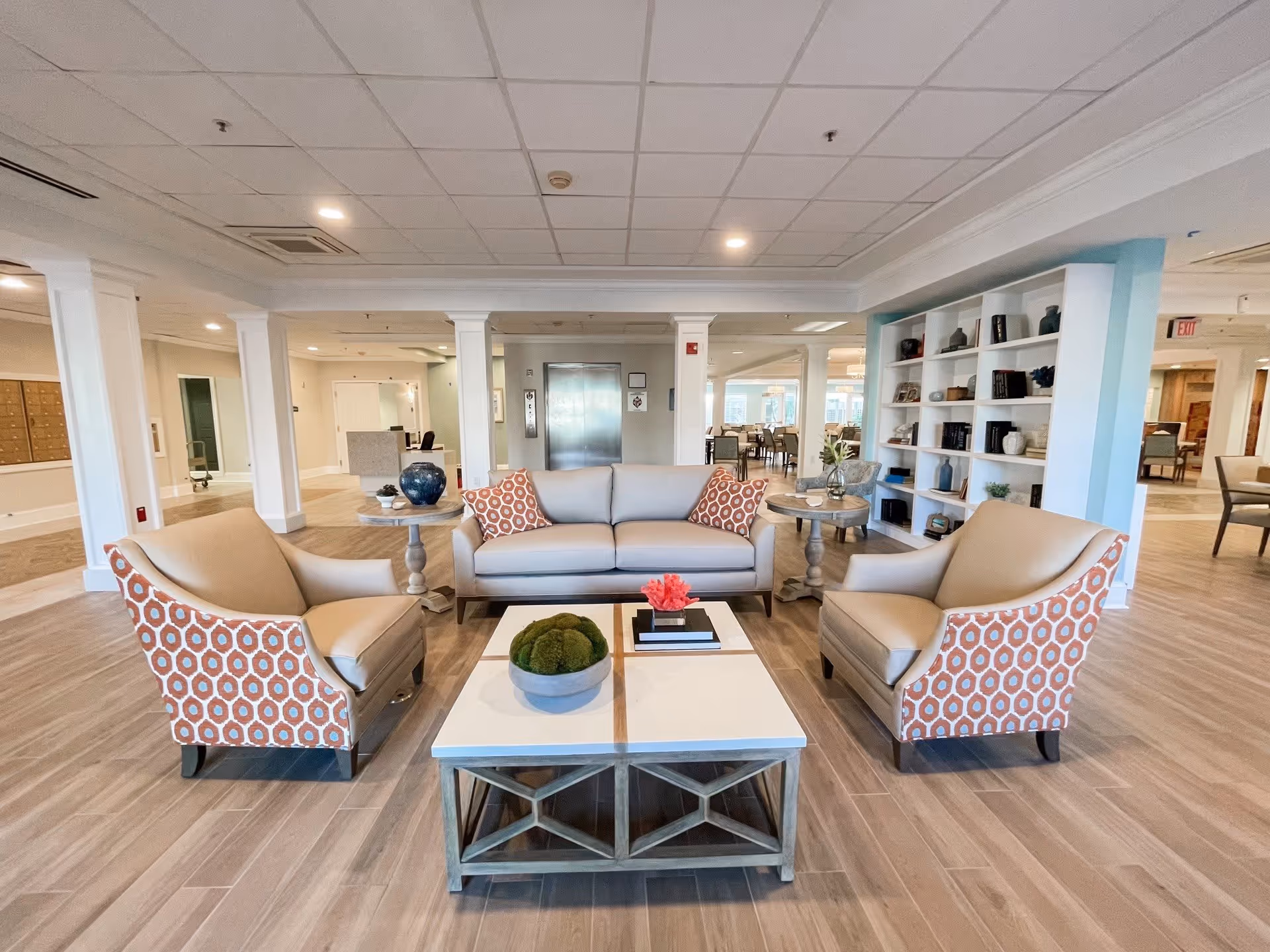 A spacious and well-lit common area with a beige sofa and two armchairs featuring orange and white patterned backs. In front of the seating is a modern coffee table with decorative items including a bowl with green moss and a small pink flower. The room has wooden flooring, white pillars, a bookshelf with various decorative items and books, and an elevator in the background. The area appears clean and inviting, likely a lounge or waiting area in a senior living facility.