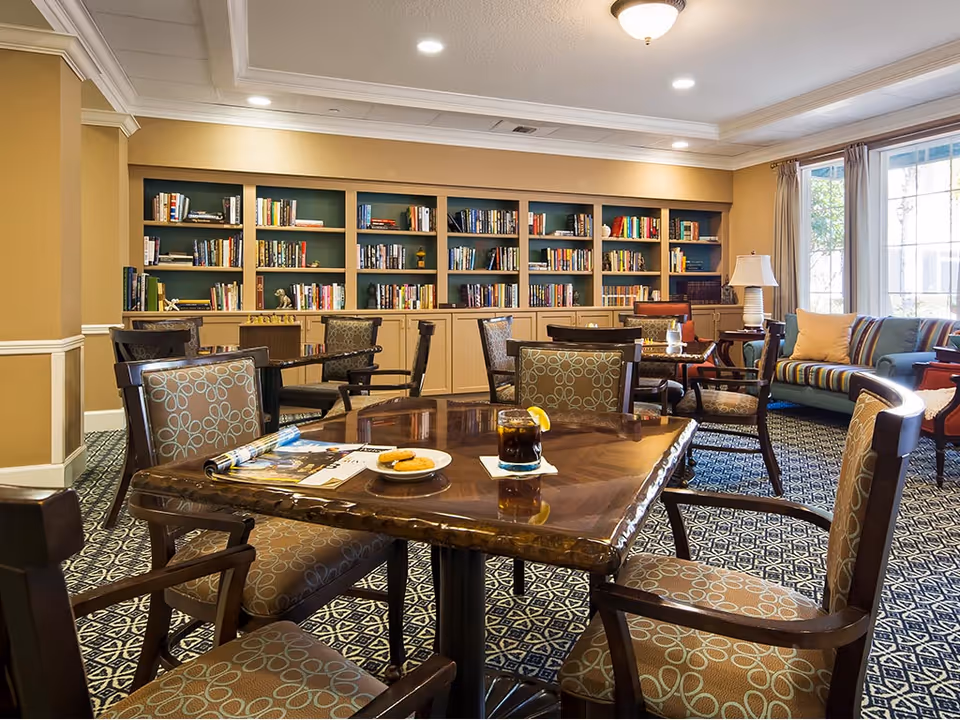 A cozy common area with a wooden table and four cushioned chairs in the foreground. On the table are a glass of dark beverage with a lemon slice, a plate with cookies, and some magazines. In the background, there is a large built-in bookshelf filled with books, a striped sofa with cushions, a side table with a lamp, and large windows with curtains letting in natural light.