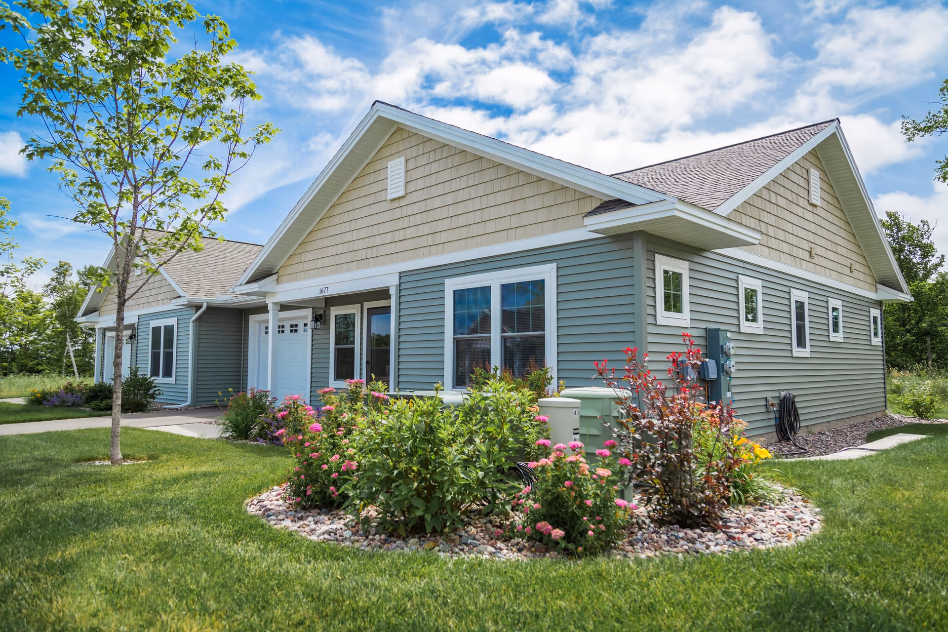 Front exterior of a single-story senior living unit with green siding, white trim, and landscaped flower beds under a blue sky.