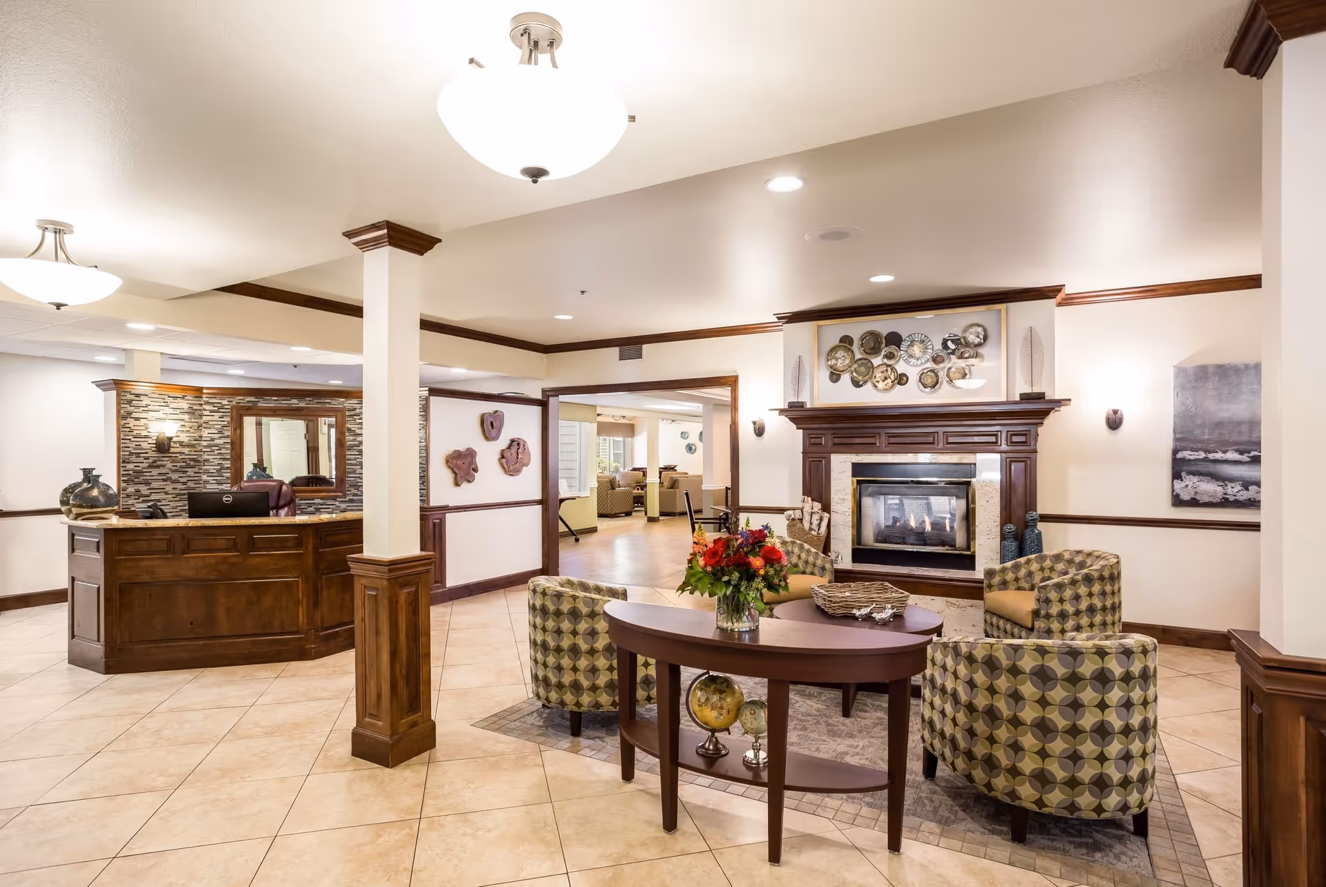A cozy senior living facility common area with a wooden reception desk on the left, a seating area with patterned armchairs around a wooden table with a flower vase in the center, and a fireplace with decorative plates above it on the right. The room has tiled floors, cream-colored walls with wooden trim, and ceiling lights.