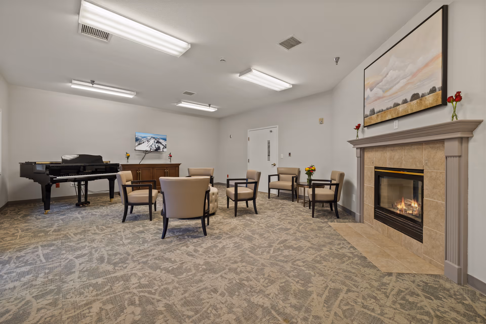 Well-lit common room with chairs arranged around a fireplace, a piano, and wall art.