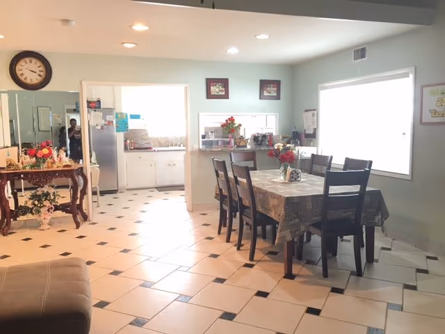 A dining room with a table set for meals and a kitchen area in the background.