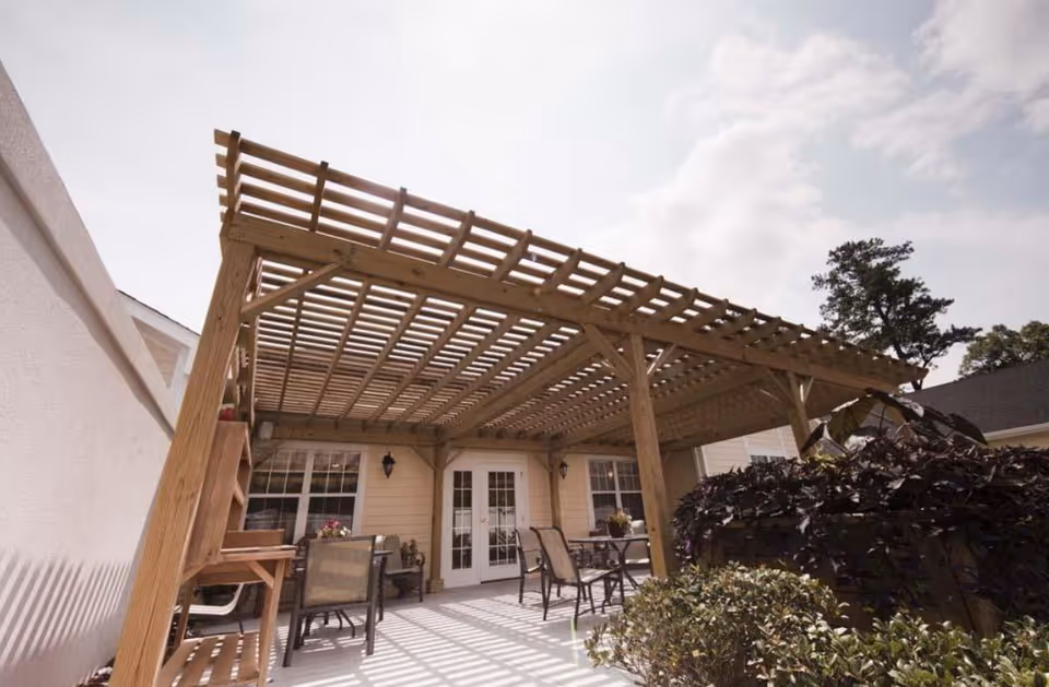 Outdoor patio area with a wooden pergola shading a seating and dining set in front of building doors and windows.