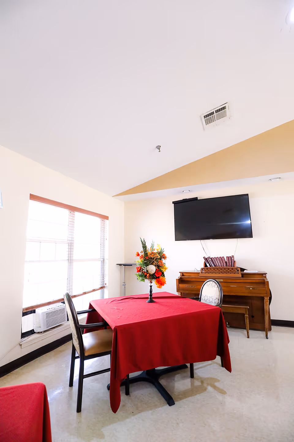 A small dining area with a table covered in a red tablecloth and two chairs. A vase with a colorful flower arrangement is placed on the table. A piano with a bench is against the wall, and a flat-screen TV is mounted above it. Large windows with blinds allow natural light into the room.