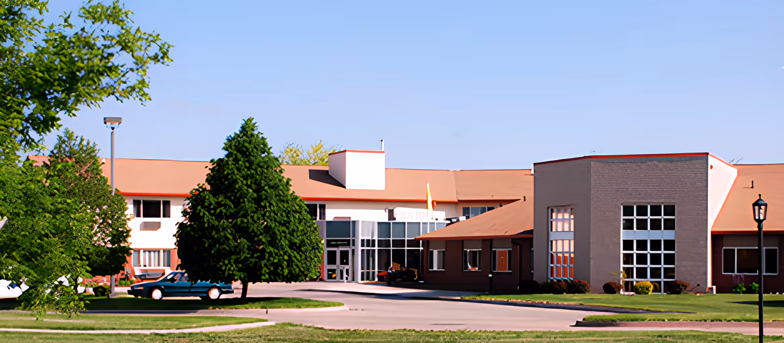 Exterior view of a single-story senior living facility building with a reddish-brown roof, large windows, and surrounded by green trees and grass under a clear blue sky.