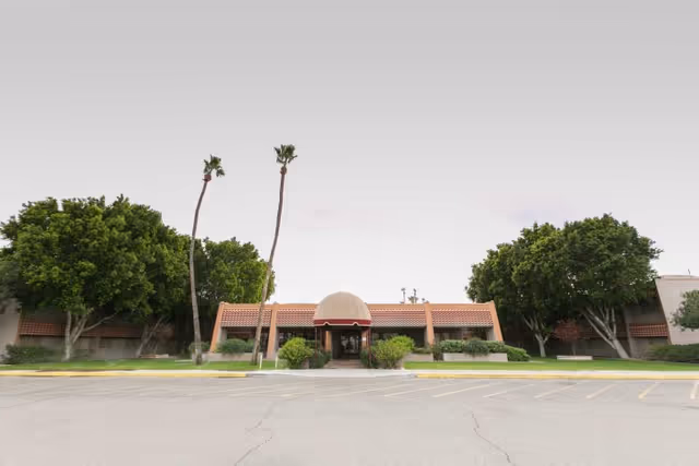 Front exterior view of a single-story building with a dome-shaped entrance canopy, surrounded by green trees and a parking lot in front under a clear sky.
