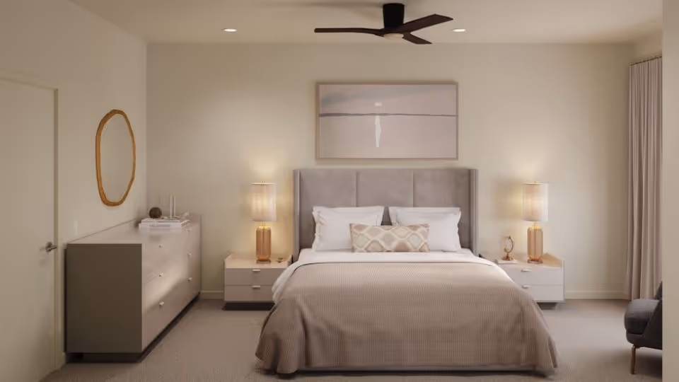A neatly arranged bedroom with a large bed centered against the wall, featuring a padded headboard, white pillows, and a beige bedspread. On each side of the bed are matching nightstands with table lamps emitting warm light. Above the bed hangs a minimalist framed artwork. To the left is a dresser with a round mirror above it, and to the right is a window with beige curtains. The room has neutral tones and a ceiling fan with dark blades.