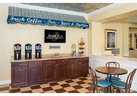 A small bistro area with a countertop holding three beverage dispensers, a glass container with pastries, and a tiered stand with fruit. Above the counter is a striped awning with the text 'Fresh Coffee - Tea - Juice & Pastries' and a sign that reads 'Grand Villa Senior Living Community'. There is a round table with four chairs with teal cushions in the foreground. The floor has a checkered pattern and the walls are painted light yellow with white trim.