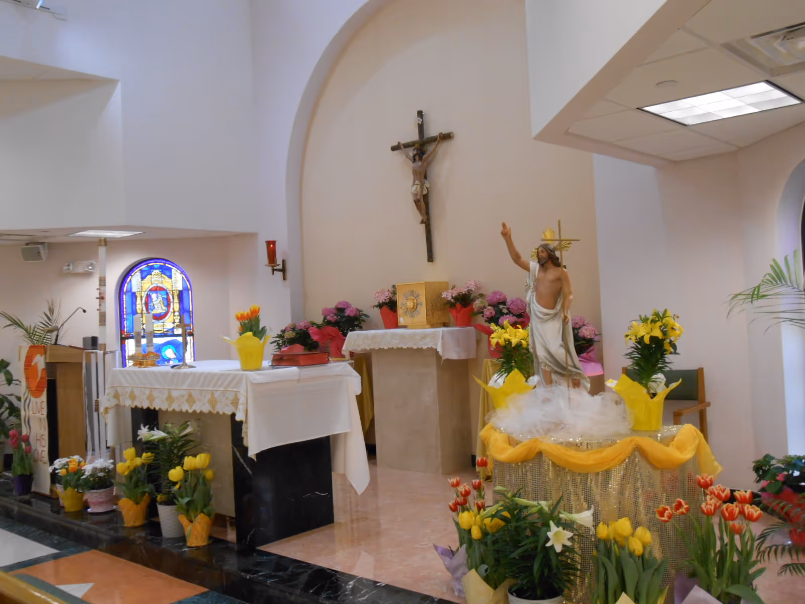Interior view of a chapel with religious decorations including a crucifix on the wall, a statue of Jesus, an altar covered with a white cloth, various flower arrangements, and a stained glass window in the background.
