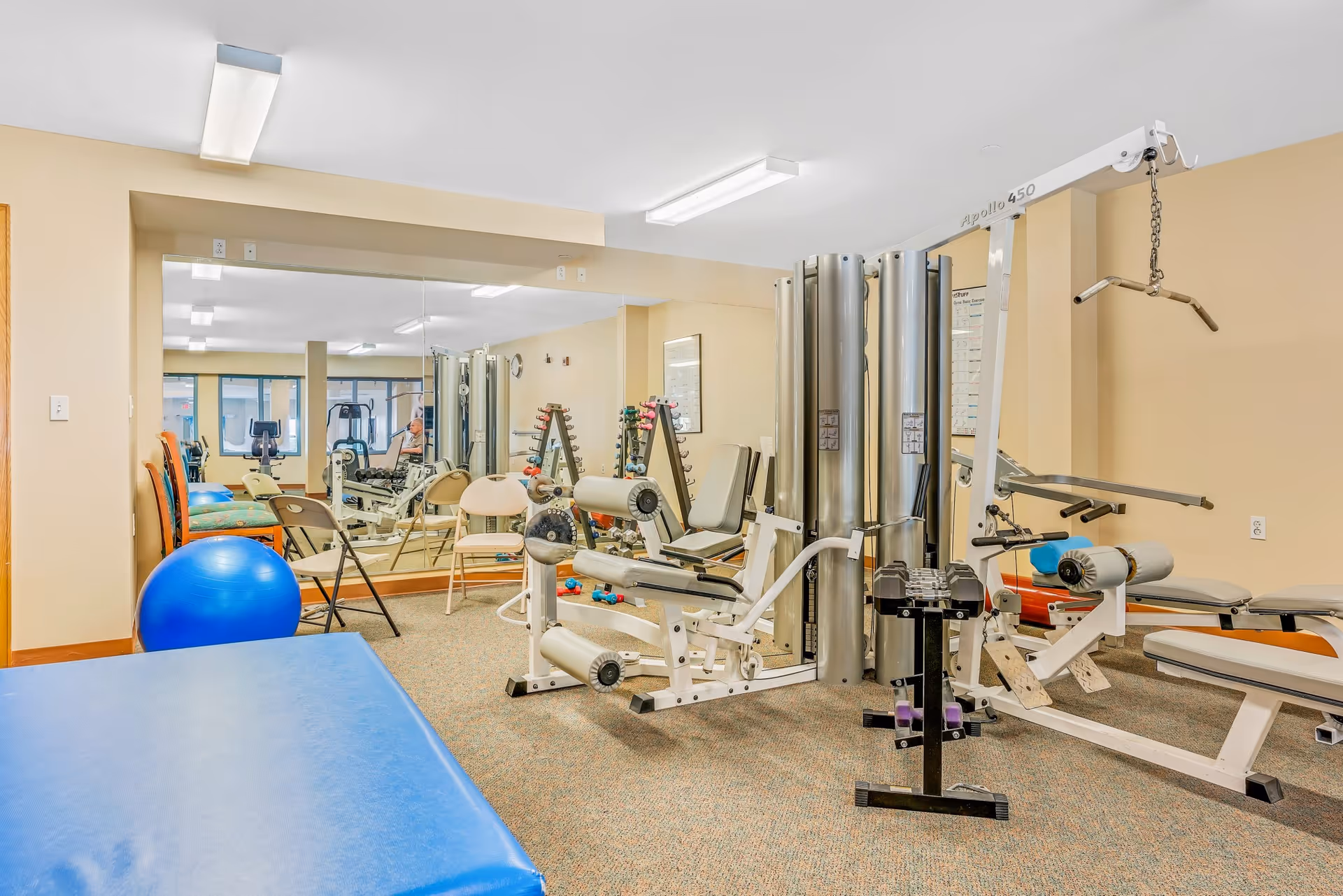 A fitness room with various exercise equipment including a multi-station weight machine, dumbbells on a rack, a blue exercise ball, and a padded blue mat. The room has beige walls, carpeted floor, and a large mirror on one wall reflecting the equipment and chairs.