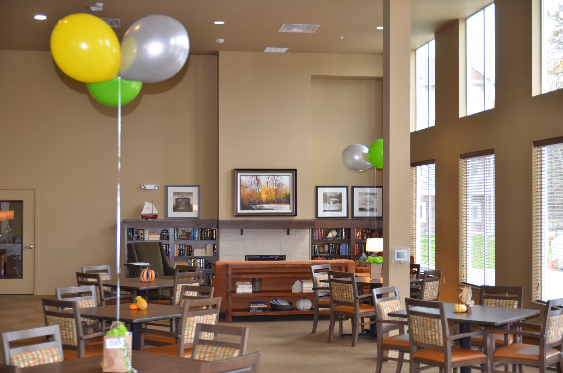 A spacious and well-lit common area with multiple tables and chairs arranged for seating. The room features large windows with blinds allowing natural light to fill the space. There are colorful balloons tied to some tables, and a fireplace with framed artwork above it is visible in the background. Bookshelves flank the fireplace, and the walls are painted in warm neutral tones.