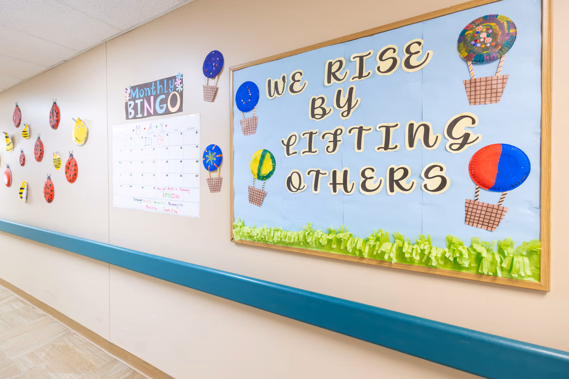 A hallway wall decorated with a bulletin board featuring the phrase 'We rise by lifting others' surrounded by colorful hot air balloon crafts. Next to it is a monthly bingo calendar and wall decorations of ladybugs and bees made from paper plates.