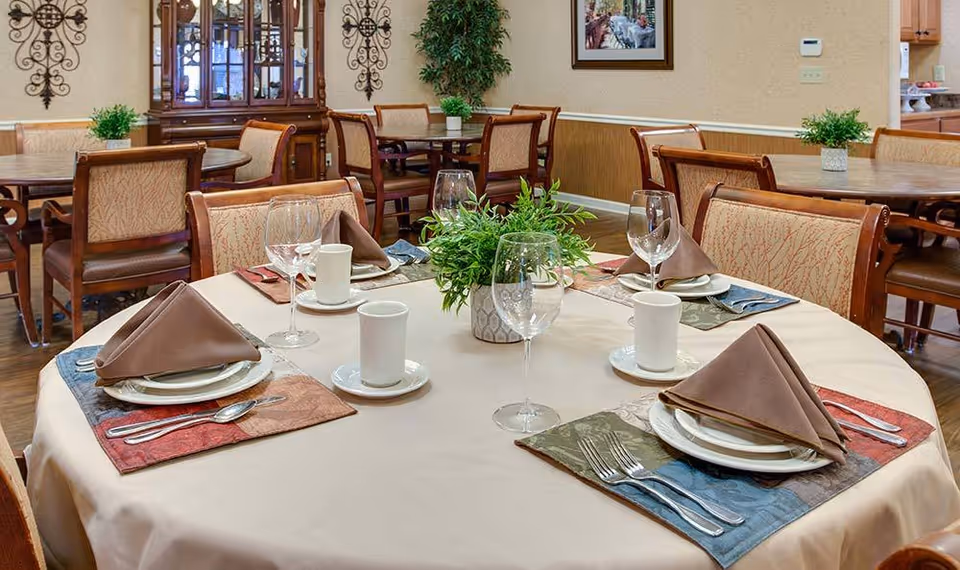 A dining room in a senior living facility with a round table set for four people. Each place setting includes a folded brown napkin, white plates, silverware, a wine glass, and a white coffee cup on a saucer. The table has a beige tablecloth and a small green plant centerpiece. In the background, there are additional tables and chairs, a wooden cabinet with glass doors, wall decorations, and a framed picture.