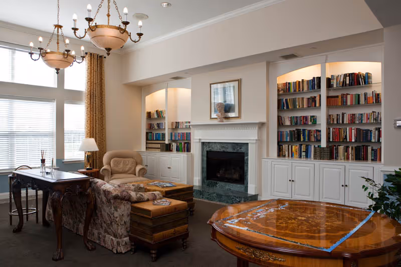A cozy living room with a floral patterned sofa, an armchair, and a wooden coffee table in front of a fireplace with a marble surround. Built-in bookshelves filled with books flank the fireplace. Large windows with white blinds and gold curtains let in natural light. Two chandeliers hang from the ceiling, and there is a wooden table with intricate designs in the foreground.