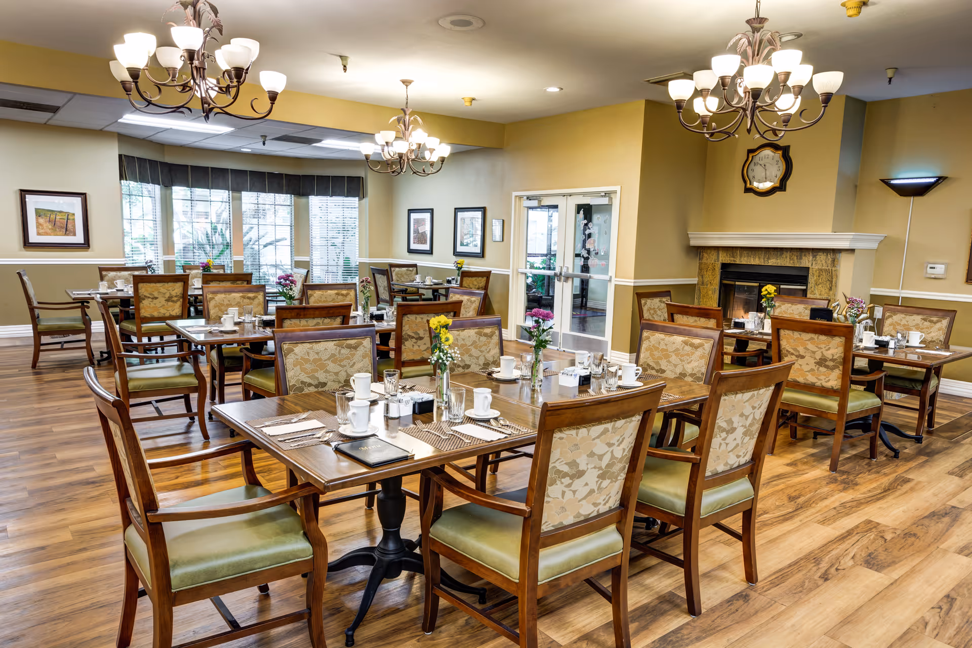 Well-lit dining room with multiple set wooden tables and chairs, chandeliers, and a fireplace.