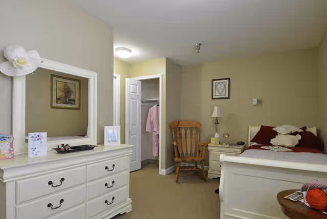 A cozy bedroom in a senior living facility with a white dresser and mirror on the left, a wooden rocking chair and nightstand with a lamp in the center, and a single bed with red and white bedding on the right. An open closet door reveals hanging clothes, and framed artwork decorates the beige walls.