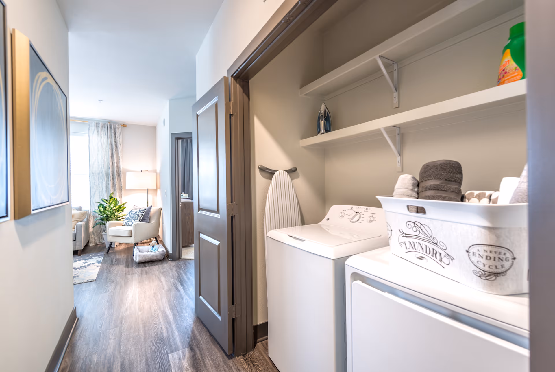 A hallway laundry closet with a washer, dryer and shelves opens onto a bright living room with an armchair and lamp.