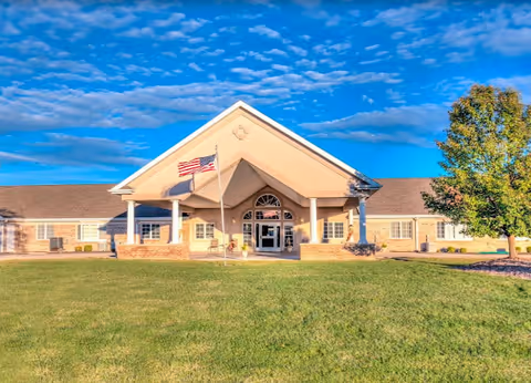Front exterior view of Brookstone Estates of Mattoon North, a single-story building with a large covered entrance supported by columns, an American flag on a flagpole in front, and a well-maintained lawn under a partly cloudy blue sky.