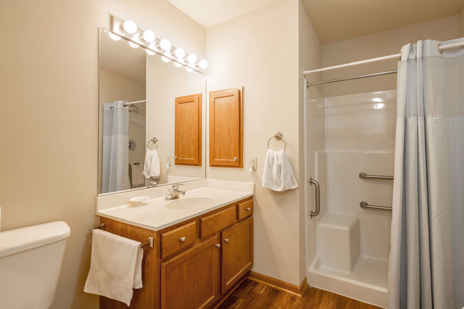 Well-lit bathroom with a sink vanity and mirror, toilet, and a shower stall with grab bars and a curtain.