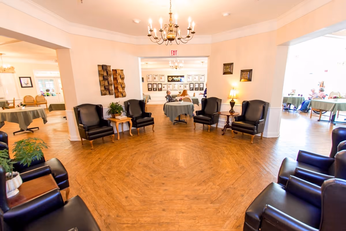 A spacious and well-lit common area in a senior living facility with multiple black leather armchairs arranged around small wooden tables. The room has wooden flooring, beige walls, and a chandelier hanging from the ceiling. In the background, there are tables covered with green tablecloths where people are seated, and framed pictures on the walls.